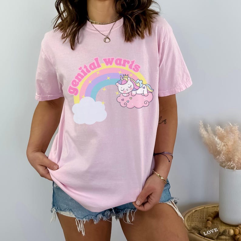 model is wearing comfort colors tee in blossom (light pink) with genital warts design on front with a rainbow stars and a unicorn