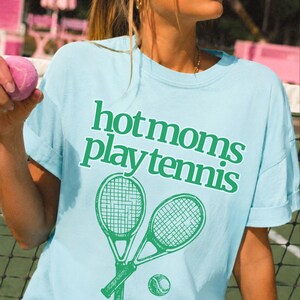 May include: A light blue crew neck t-shirt featuring the green text "hot moms play tennis" printed on the front. Below the text is a graphic illustration of two crossed tennis rackets and a single tennis ball in a matching green colour.