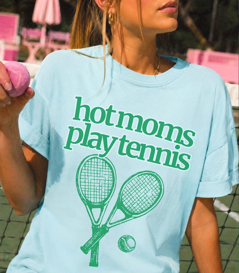 May include: A light blue crew neck t-shirt featuring the green text "hot moms play tennis" printed on the front. Below the text is a graphic illustration of two crossed tennis rackets and a single tennis ball in a matching green colour.