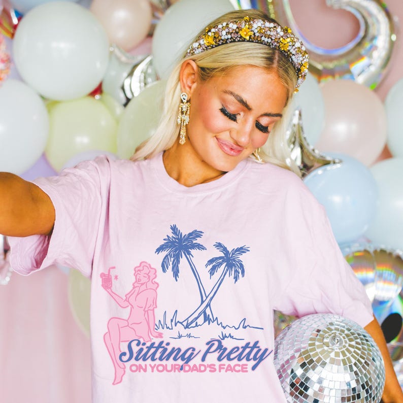 May include: A light pink t-shirt with a graphic of palm trees and a woman holding a drink. The text on the shirt reads "Sitting Pretty on Your Dad's Face". The image also includes a disco ball.