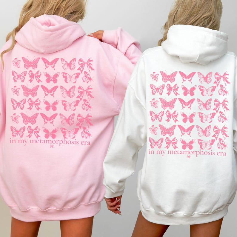 Puede incluir: Dos sudaderas con capucha, una rosa y otra blanca, presentan un dise&ntilde;o de mariposas y lazos rosas en la espalda. El texto "in my metamorphosis era" est&aacute; impreso debajo. Las sudaderas son ropa casual.
