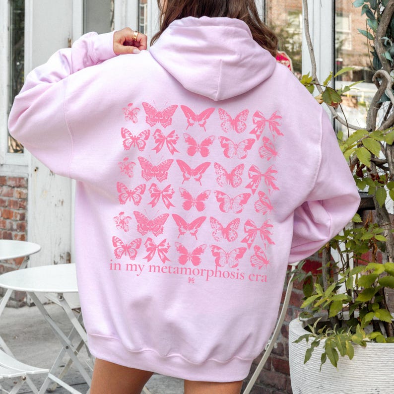 Puede incluir: Una sudadera con capucha rosa claro con capucha y mangas largas. La parte trasera de la sudadera presenta un dise&ntilde;o de mariposas y lazos rosas con el texto "in my metamorphosis era". La sudadera est&aacute; hecha de un material suave.