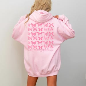 Puede incluir: Sudadera con capucha rosa claro con un dise&ntilde;o de mariposas y lazos en la espalda. El dise&ntilde;o es en un tono rosa m&aacute;s oscuro e incluye el texto "in my metamorphosis era". La sudadera tiene capucha y mangas largas.