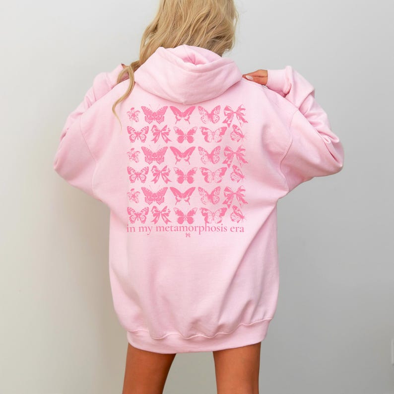 Puede incluir: Sudadera con capucha rosa claro con un dise&ntilde;o de mariposas y lazos en la espalda. El dise&ntilde;o es en un tono rosa m&aacute;s oscuro e incluye el texto "in my metamorphosis era". La sudadera tiene capucha y mangas largas.