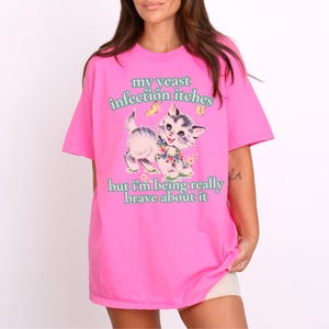 May include: A bright pink t-shirt featuring a vintage-style illustration of a kitten surrounded by flowers and butterflies. The text on the shirt reads "my yeast infection itches" and "but I'm being really brave about it."