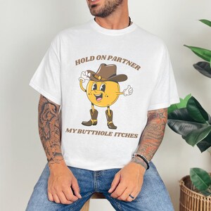 Model is wearing a gildan tee shirt in white with a smiling cowboy giving a thumbs up and it says hold on partner my butthole itches on front in brown