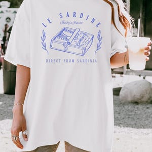 model is wearing comfort colors oversized tee in white with le sardine can and italy's finest direct from sardinia in blue on front