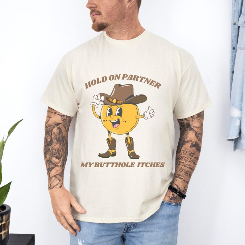 Model is wearing a gildan tee shirt in natural with a smiling cowboy giving a thumbs up and it says hold on partner my butthole itches on front in brown