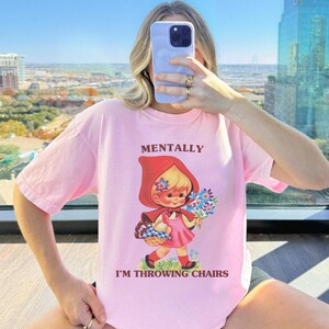 May include: A light pink t-shirt with the words "MENTALLY I'M THROWING CHAIRS" printed on the front. The graphic is a vintage-style illustration of a girl in a red hooded cape holding flowers and a basket. The shirt is a relaxed fit.