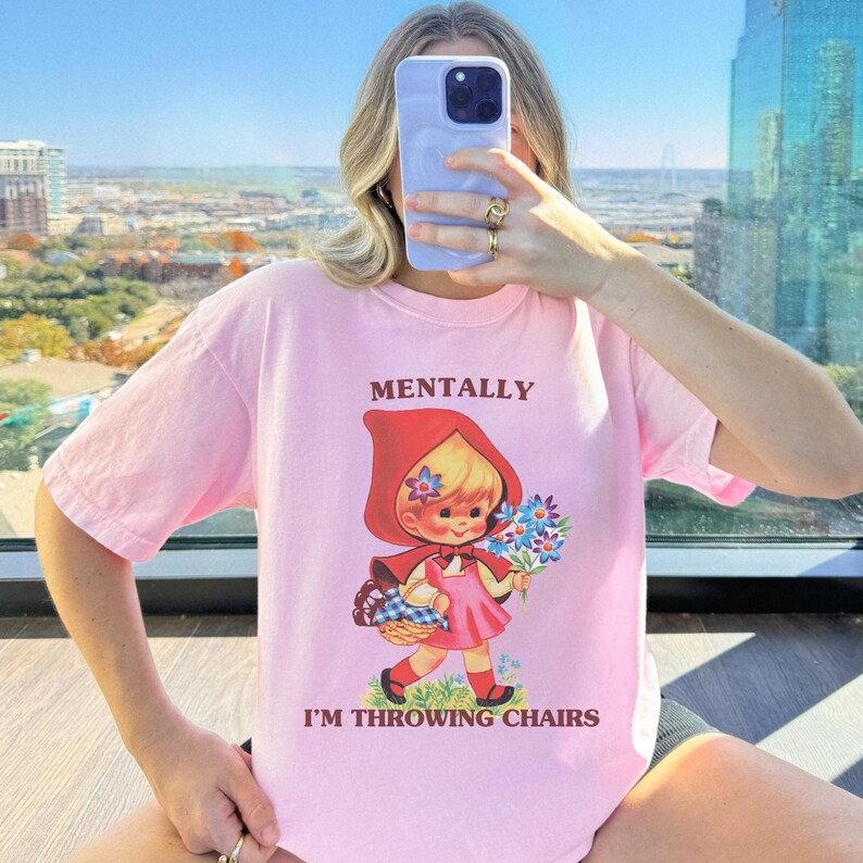 May include: A light pink t-shirt with the words "MENTALLY I'M THROWING CHAIRS" printed on the front. The graphic is a vintage-style illustration of a girl in a red hooded cape holding flowers and a basket. The shirt is a relaxed fit.