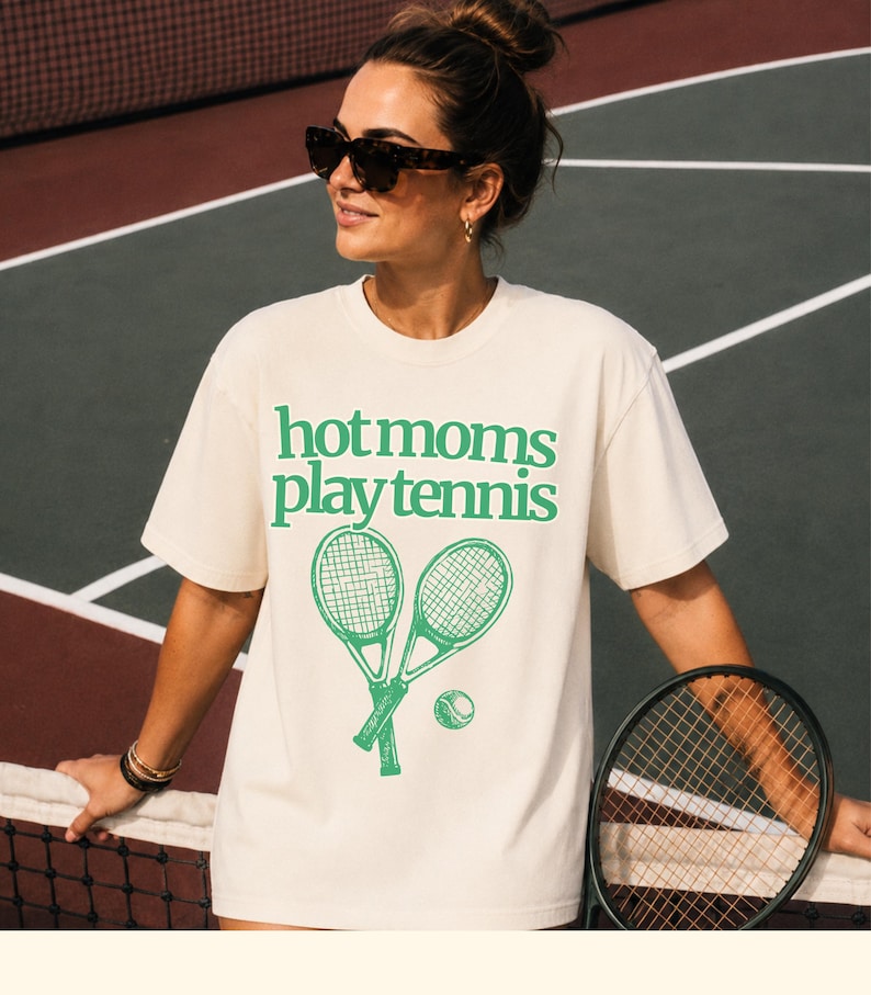 May include: A cream-coloured short-sleeve t-shirt featuring the green text "hot moms play tennis" printed on the chest, accompanied by a graphic illustration of two crossed tennis rackets and a single tennis ball positioned below the text.