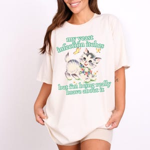 May include: Cream-colored t-shirt with a graphic of a kitten surrounded by flowers and butterflies. The text "my yeast infection itches but I'm being really brave about it" is printed in green.