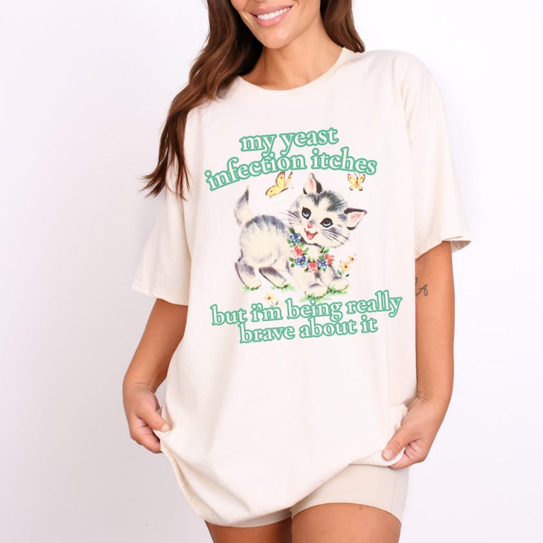 May include: Cream-colored t-shirt with a graphic of a kitten surrounded by flowers and butterflies. The text "my yeast infection itches but I'm being really brave about it" is printed in green.