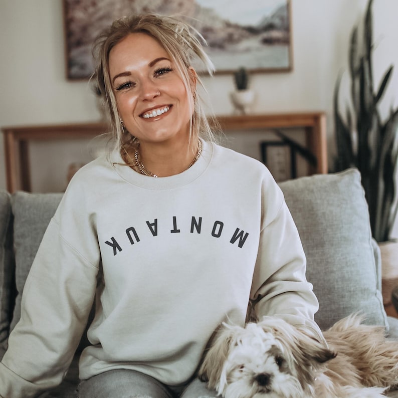 model is wearing gildan oversized crewneck sweatshirt in sand with upside down montauk in black across front