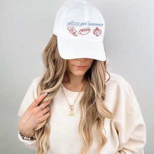 May include: White baseball cap with the text "glizzy girl summer" in blue script. The cap features red illustrations of condiments, a hot dog, and an onion. The cap is worn by a person.