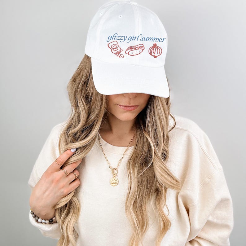 May include: White baseball cap with the text "glizzy girl summer" in blue script. The cap features red illustrations of condiments, a hot dog, and an onion. The cap is worn by a person.