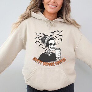 Op de afbeelding: Een crèmekleurige hoodie met een skelet dat een hoofdband draagt en koffie drinkt. De tekst "Batty Before Coffee" is op de hoodie gedrukt.