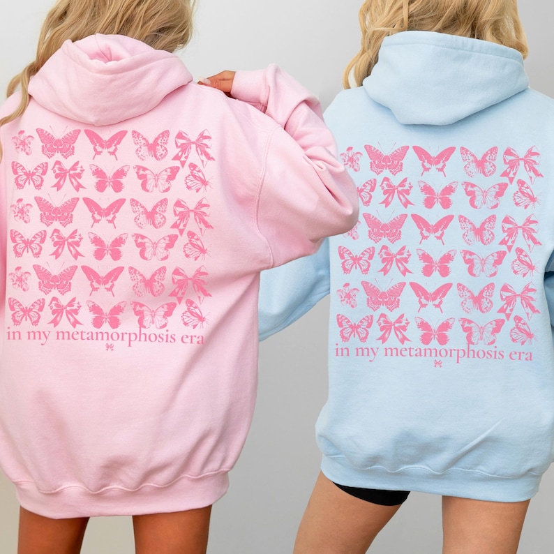 Puede incluir: Dos sudaderas con capucha, una rosa claro y otra azul claro, con un dise&ntilde;o de mariposas y lazos rosas en la espalda. El texto "in my metamorphosis era" est&aacute; impreso debajo. Ropa informal.