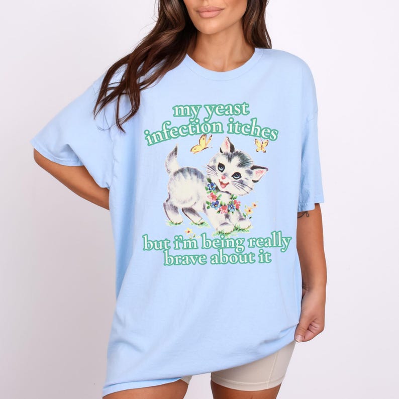 May include: Light blue t-shirt with the text "my yeast infection itches but I'm being really brave about it." The shirt features a vintage-style illustration of a gray and white kitten with a floral collar and two yellow butterflies.