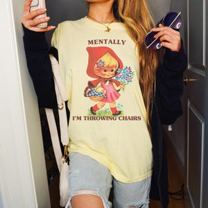 May include: A light yellow t-shirt with the text "MENTALLY I'M THROWING CHAIRS" and a graphic of a cartoon girl in a red hood holding flowers. The shirt is paired with light blue ripped jeans and a black jacket.