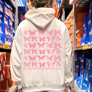 Puede incluir: Sudadera con capucha color crema con un dise&ntilde;o de mariposas rosas en la espalda. El dise&ntilde;o incluye varias ilustraciones de mariposas y el texto "in my metamorphosis era". La sudadera tiene capucha y mangas largas.