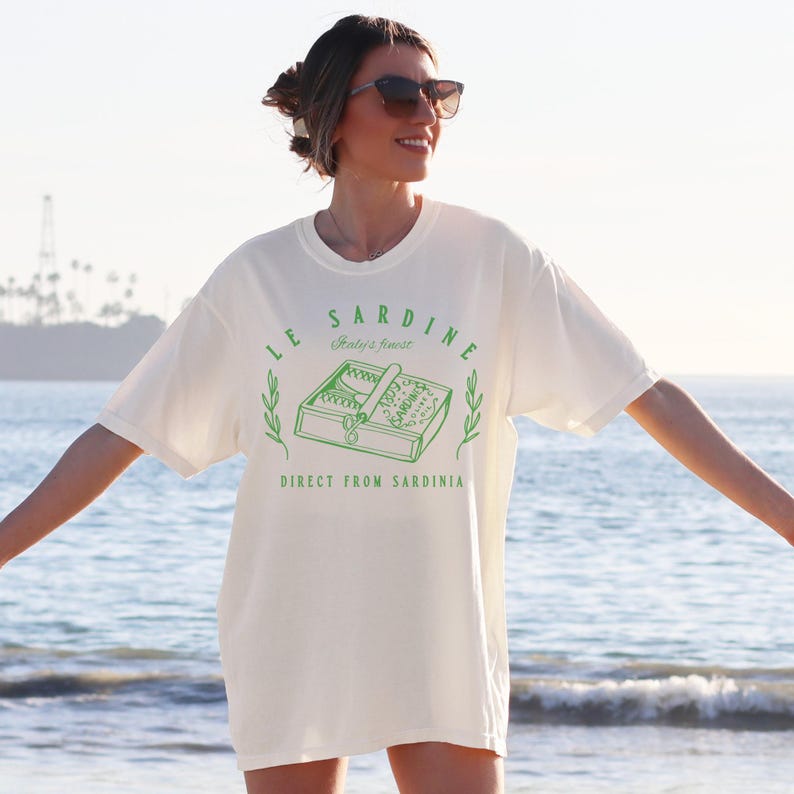 model is wearing comfort colors oversized tee in ivory (off white/natural) with le sardine can and italy's finest direct from sardinia in green on front