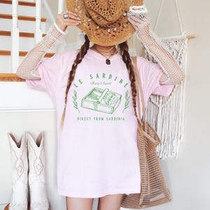 model is wearing comfort colors oversized tee in blossom (light pink) with le sardine can and italy's finest direct from sardinia in green on front