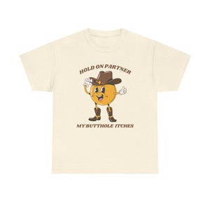 Model is wearing a gildan tee shirt in natural with a smiling cowboy giving a thumbs up and it says hold on partner my butthole itches on front in brown