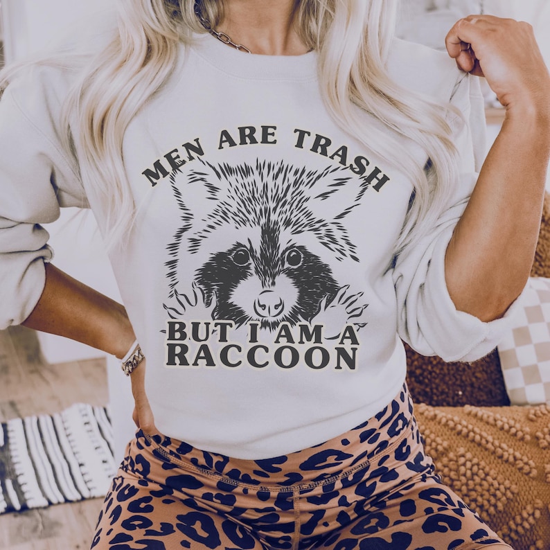 May include: A cream-coloured sweatshirt featuring a raccoon graphic and the text "MEN ARE TRASH BUT I AM A RACCOON" in black. The raccoon illustration is detailed with black and white shading. The sweatshirt is paired with leopard print leggings.