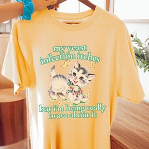 May include: Yellow t-shirt with a graphic of a kitten wearing a floral collar, with the text "my yeast infection itches but I'm being really brave about it" in green. The kitten is gray and white with a blue and white floral collar.