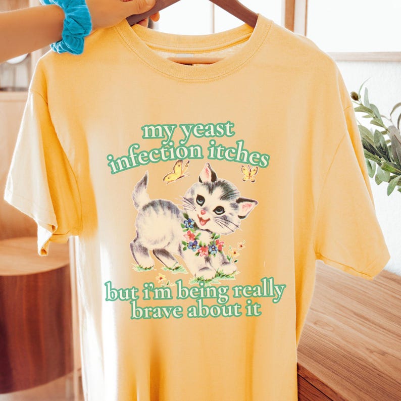 May include: Yellow t-shirt with a graphic of a kitten wearing a floral collar, with the text "my yeast infection itches but I'm being really brave about it" in green. The kitten is gray and white with a blue and white floral collar.