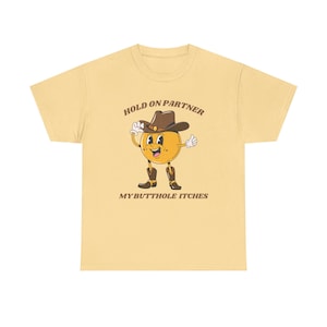 Model is wearing a gildan tee shirt in yellow haze with a smiling cowboy giving a thumbs up and it says hold on partner my butthole itches on front in brown