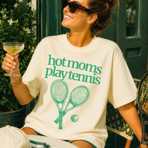 May include: A cream-coloured short-sleeve t-shirt featuring the green text "hot moms play tennis" printed above a graphic of two crossed tennis rackets and a tennis ball. The shirt has a crew neckline and a relaxed fit, showcasing a casual style.