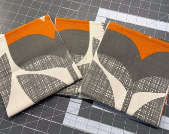 May include: Three folded fabric squares with a modern design. The fabric features a pattern of grey, white, and orange shapes. The design includes a combination of solid colours and a textured, cross-hatched pattern. The squares are neatly stacked.