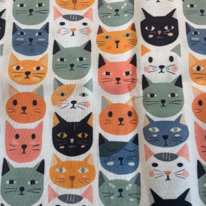 May include: A white fabric featuring a repeating pattern of stylized cat faces in various colors including orange, blue, pink, black, and sage green. Each cat face displays simple line-drawn features, including eyes, whiskers, and small noses, arranged in a grid-like layout across the textile surface.