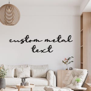 May include: Black metal wall art spelling out "custom metal text" in a cursive font. The text is mounted on a white wall above a sofa and other furniture. The room is decorated in neutral tones.