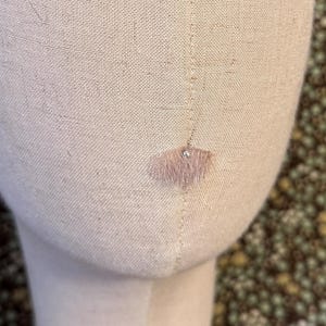 May include: A beige fabric mannequin head displaying a small, delicate, fan-shaped earring with a light pink fringe and a single clear crystal accent positioned near the center of the head form.
