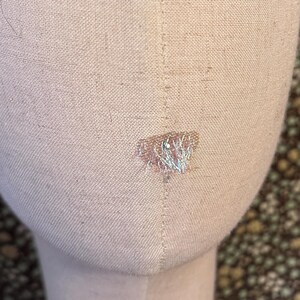 May include: A beige fabric-covered mannequin head with a vertical seam running down the center and a small, light-colored embroidered detail featuring thin, silver-toned threads located on the lower front portion of the oval-shaped form.
