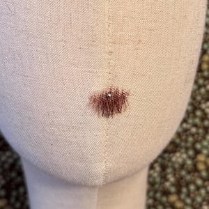 May include: A beige fabric-covered mannequin head with a vertical seam running down the center. A small, singular patch of brown hair is attached to the lower middle section of the face, featuring a tiny silver pin placed directly in the center.