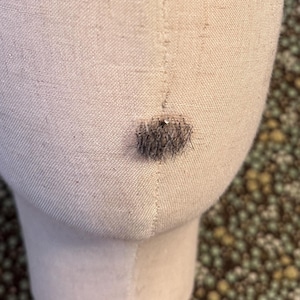 May include: A beige fabric-covered mannequin head with a smooth, oval shape and a vertical seam running down the center. A small, circular patch of dark, textured hair is attached to the lower front section of the mannequin head.