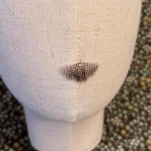 May include: A beige fabric-covered mannequin head featuring a small, dark brown, triangular-shaped patch of hair or fiber attached to the lower center area, secured with a single small silver-colored pin at the top of the patch.
