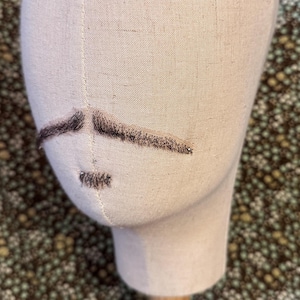 May include: A beige fabric mannequin head featuring a dark brown faux hair mustache and a small matching soul patch. The mannequin is mounted on a light-colored wooden stand, displaying the facial hair pieces clearly on the smooth textile surface.