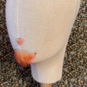May include: A beige fabric mannequin head displaying a small, orange-colored faux hair mustache and goatee set. The facial hair pieces are attached to the center of the mannequin face, featuring vibrant orange fibers with subtle yellow highlights.