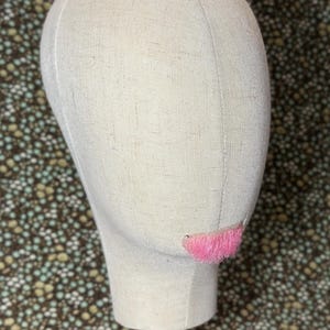 May include: A beige fabric mannequin head with a pink fringe detail, mounted on a wooden stand. The head is oval-shaped and has a smooth, textured surface. The pink fringe is located where the mouth would be.