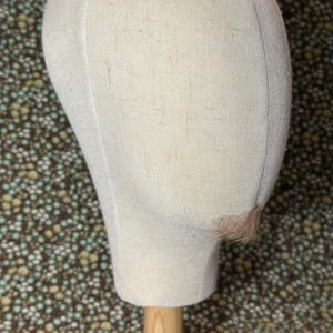May include: A cream-colored wig head form made of fabric, mounted on a wooden stand. The head form has a smooth, rounded shape, with a small section of hair attached near the ear. The background is a floral pattern.