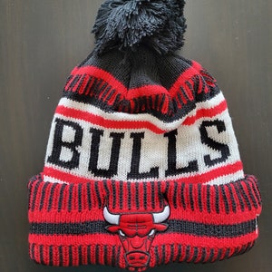 May include: A knitted beanie in black, red, and white, featuring the word "BULLS" in bold white letters. The hat has a black pom-pom on top and a red bull logo on the front. The hat is designed for cold weather.