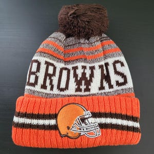 May include: An orange, white, and brown knit beanie with the word "BROWNS" in brown block letters. The beanie features a brown pom-pom and a patch of a football helmet.