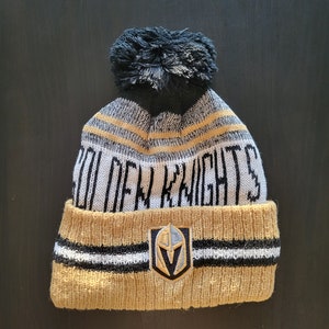 May include: A knitted beanie in gold, black, and white, featuring the text "GOLDEN KNIGHTS" and a team logo. The hat has a black pom-pom on top and is designed for cold weather.