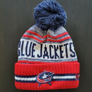 May include: A knitted beanie in red, white, and blue, featuring the text "BLUE JACKETS" across the crown. The hat has a navy blue pom-pom and a team logo patch on the folded brim. A winter accessory.