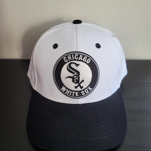 May include: A white and black baseball cap with the Chicago White Sox logo. The cap has a white crown, a black peak, and a black button. The logo is a circular emblem with the team name and logo.